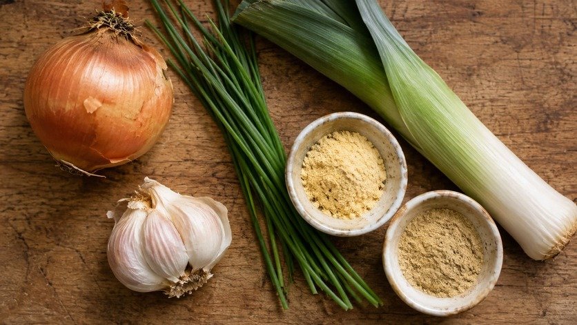 Whole onions, garlic bulbs, chives, leeks, and small bowls of onion and garlic powder on a wooden board